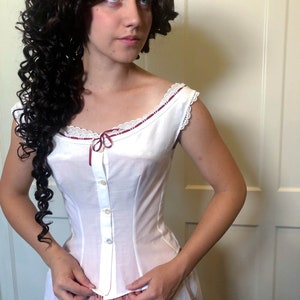 May include: White cotton corset with a scoop neckline, adorned with lace trim and a burgundy ribbon. The corset features a button-down front and a fitted silhouette, suggesting a vintage or historical style. The garment is part of a two-piece set.