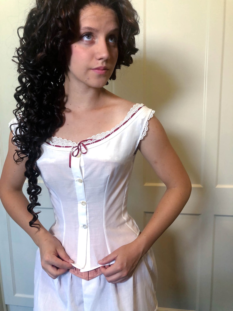 May include: White cotton corset with a scoop neckline, adorned with lace trim and a burgundy ribbon. The corset features a button-down front and a fitted silhouette, suggesting a vintage or historical style. The garment is part of a two-piece set.