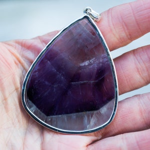 May include: A large, teardrop-shaped pendant with a polished, deep purple amethyst stone. The gemstone is set in a silver-colored metal frame with a loop for a chain. The pendant is held in a hand.