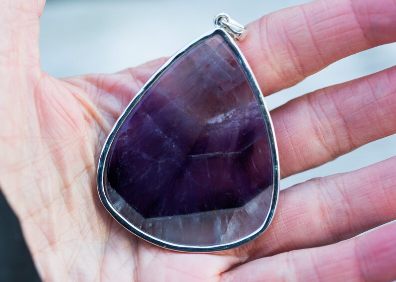 May include: A large, teardrop-shaped pendant with a polished, deep purple amethyst stone. The gemstone is set in a silver-colored metal frame with a loop for a chain. The pendant is held in a hand.