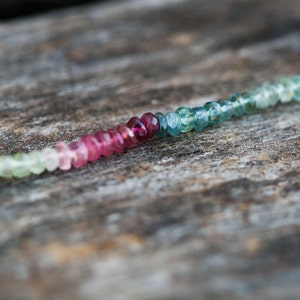 May include: A strand of faceted gemstone beads in shades of green, pink, and blue. The beads are strung on a clear thread and are arranged in a gradient pattern.