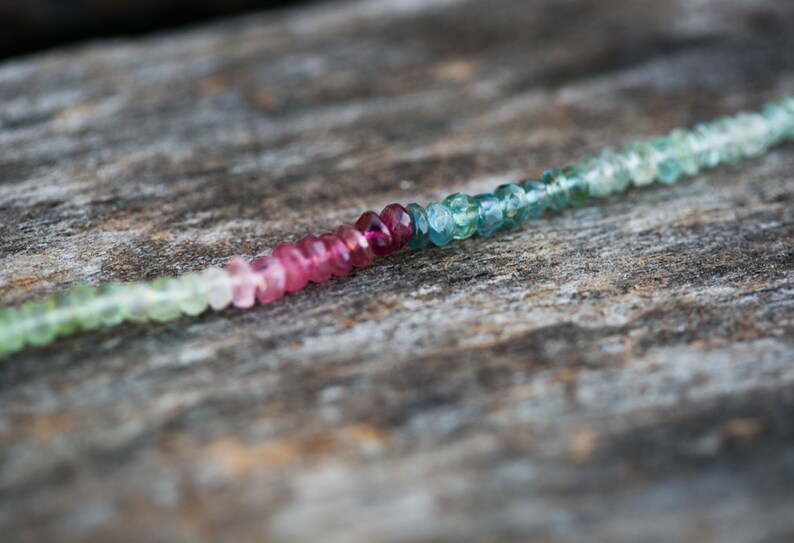 May include: A strand of faceted gemstone beads in shades of green, pink, and blue. The beads are strung on a clear thread and are arranged in a gradient pattern.