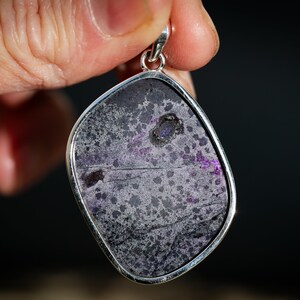 May include: A pendant with a silver-colored frame, showcasing a stone with a mottled pattern of gray, black, and purple hues. The stone has a unique, irregular shape, and the pendant is held up by a hand against a dark background.