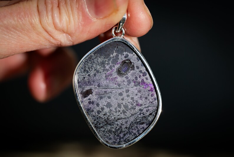May include: A pendant with a silver-colored frame, showcasing a stone with a mottled pattern of gray, black, and purple hues. The stone has a unique, irregular shape, and the pendant is held up by a hand against a dark background.