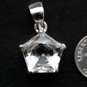 May include: A pentagonal clear gemstone pendant set in a silver-toned metal prong setting with a bail at the top. The pendant is positioned next to a United States dime for scale, showing the relative size of the faceted stone and its metal mount.