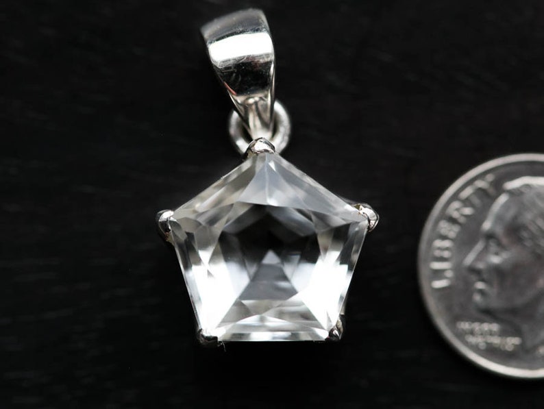 May include: A pentagonal clear gemstone pendant set in a silver-toned metal prong setting with a bail at the top. The pendant is positioned next to a United States dime for scale, showing the relative size of the faceted stone and its metal mount.