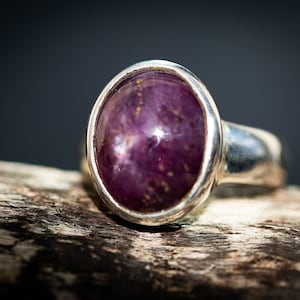 May include: A silver ring with a large, oval-shaped, deep purple gemstone. The gemstone has a star-like effect and is set in a polished silver band. The ring is displayed on a piece of weathered wood, with a dark background.