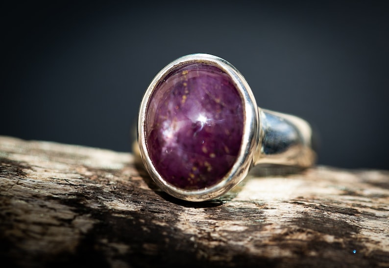 May include: A silver ring with a large, oval-shaped, deep purple gemstone. The gemstone has a star-like effect and is set in a polished silver band. The ring is displayed on a piece of weathered wood, with a dark background.