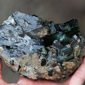 May include: A rough gemstone with dark green and black crystalline formations. The mineral is held in a hand, highlighting its natural, unpolished texture and colour variations. The stone has a dark, reflective surface.