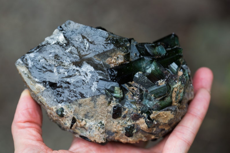 May include: A rough gemstone with dark green and black crystalline formations. The mineral is held in a hand, highlighting its natural, unpolished texture and colour variations. The stone has a dark, reflective surface.