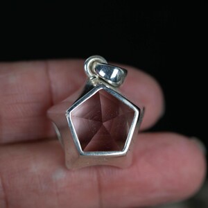 May include: A pentagonal pendant featuring a faceted, translucent, light pink gemstone set within a polished silver-toned metal frame. The pendant includes a small, integrated silver-toned metal bail at the top for attachment to a chain or cord.