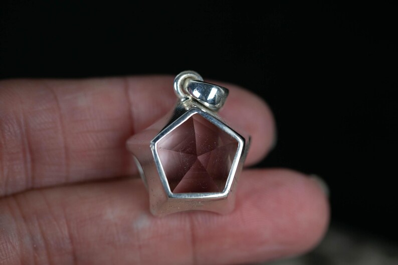 May include: A pentagonal pendant featuring a faceted, translucent, light pink gemstone set within a polished silver-toned metal frame. The pendant includes a small, integrated silver-toned metal bail at the top for attachment to a chain or cord.