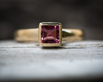 May include: A gold ring with a square-shaped pink gemstone.