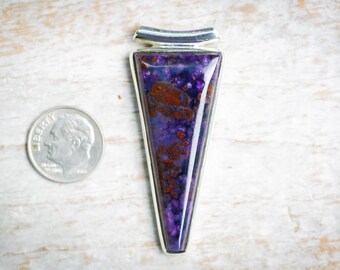 May include: A triangular pendant with a vibrant purple and red stone set in a silver-colored metal frame. The pendant is next to a U.S. dime coin for size comparison. The stone has a speckled appearance.