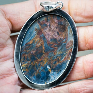 May include: Oval pendant with a silver-colored frame. The pendant features a swirling pattern of blue, brown, and gold colors. The pendant is held in a hand, showcasing the jewelry's size and design.