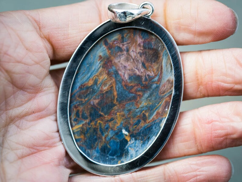 May include: Oval pendant with a silver-colored frame. The pendant features a swirling pattern of blue, brown, and gold colors. The pendant is held in a hand, showcasing the jewelry's size and design.