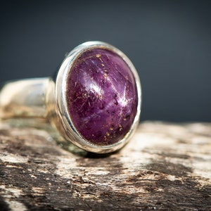 May include: A silver ring featuring a large, oval-shaped, deep purple gemstone. The gemstone has a star-like effect and is set in a polished silver band. The ring is displayed on a piece of weathered wood.