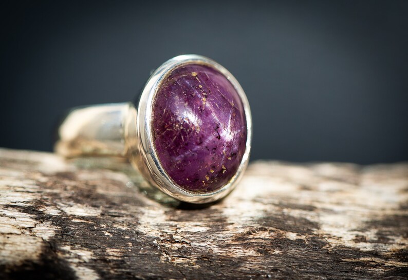May include: A silver ring featuring a large, oval-shaped, deep purple gemstone. The gemstone has a star-like effect and is set in a polished silver band. The ring is displayed on a piece of weathered wood.
