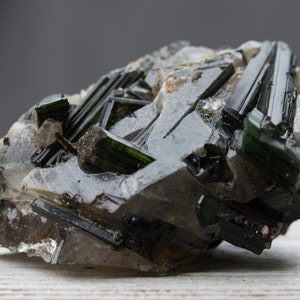 May include: A close-up of a raw mineral specimen. The stone features dark green, elongated crystals embedded in a matrix of clear and grey quartz. The crystals have a glassy lustre, and the overall appearance is rough and natural.
