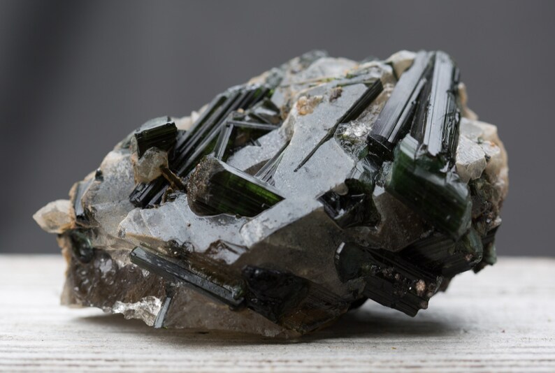 May include: A close-up of a raw mineral specimen. The stone features dark green, elongated crystals embedded in a matrix of clear and grey quartz. The crystals have a glassy lustre, and the overall appearance is rough and natural.