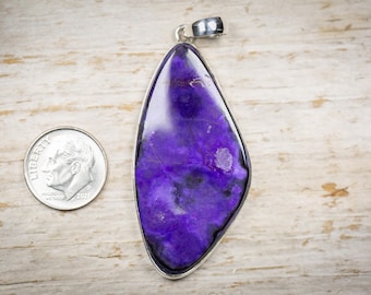 May include: A pendant with a large, polished, purple stone set in a silver-colored metal frame. The stone has a marbled appearance with dark purple and white inclusions. A dime is next to the pendant for size comparison.
