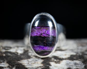 May include: A silver ring with an oval stone, displaying layers of black and purple. The stone is set in a polished silver band. The ring is shown against a dark background, highlighting the colours and textures of the stone.