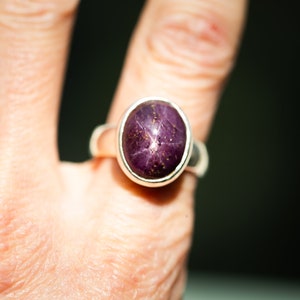 May include: Oval-shaped ring featuring a deep purple gemstone with a star-like pattern. The gemstone is set in a silver band. The ring is shown on a finger, highlighting the jewelry's design and color.