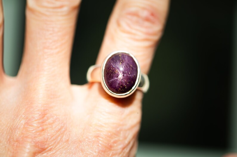 May include: Oval-shaped ring featuring a deep purple gemstone with a star-like pattern. The gemstone is set in a silver band. The ring is shown on a finger, highlighting the jewelry's design and color.