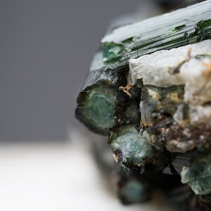 May include: Close-up of a raw gemstone with a deep green colour. The mineral displays crystalline structures and a rough, textured surface. White and tan mineral deposits are also visible.