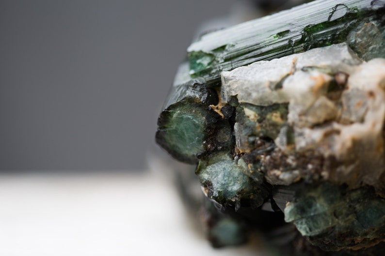 May include: Close-up of a raw gemstone with a deep green colour. The mineral displays crystalline structures and a rough, textured surface. White and tan mineral deposits are also visible.