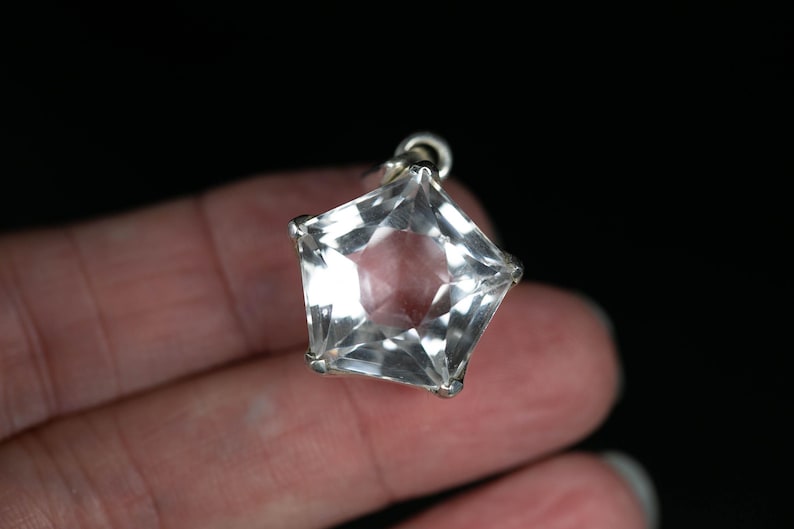 May include: A clear, faceted, star-shaped pendant set in a silver-toned metal frame. The pendant features multiple triangular facets that catch the light, creating a brilliant, reflective surface. It includes a small metal loop at the top for attachment.