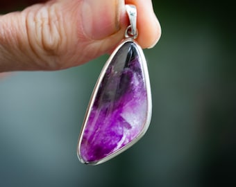 May include: A teardrop-shaped pendant with a vibrant purple and black stone set in a silver-colored metal frame. The stone has a marbled appearance with shades of purple and black. The pendant is held up by a silver bail.