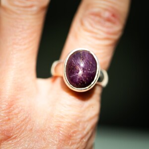May include: A silver ring featuring a large, oval-cut, purplish-red gemstone with a star-like pattern. The ring is set in a simple, polished silver band. The gemstone's color and star effect are prominent.