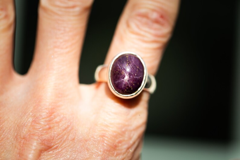 May include: A silver ring featuring a large, oval-cut, purplish-red gemstone with a star-like pattern. The ring is set in a simple, polished silver band. The gemstone's color and star effect are prominent.