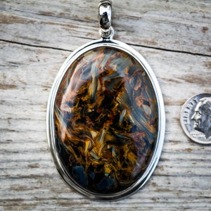 May include: Oval pendant with a polished gemstone, featuring swirling patterns of brown, gold, and blue, set in a silver frame. A dime is placed next to the pendant for size comparison.
