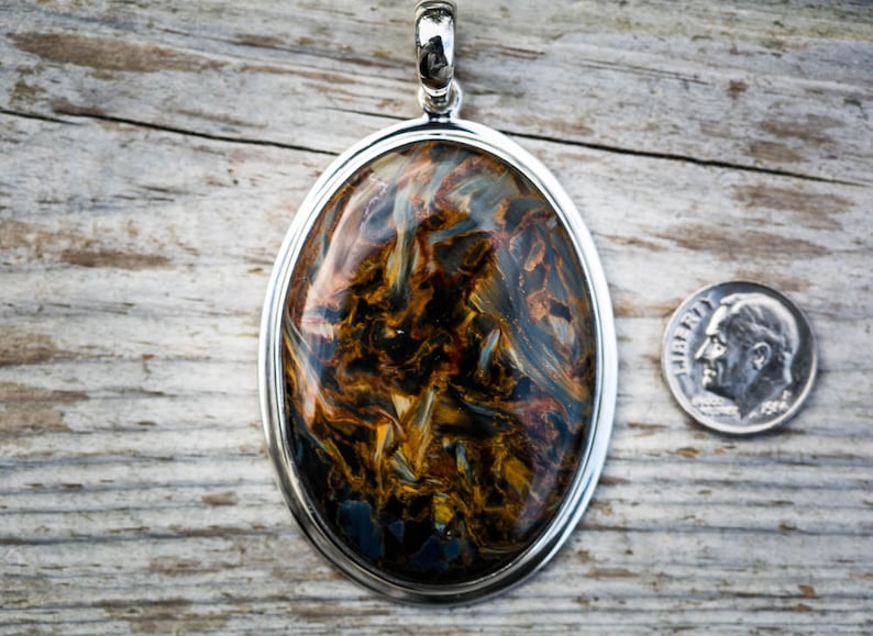 May include: Oval pendant with a polished gemstone, featuring swirling patterns of brown, gold, and blue, set in a silver frame. A dime is placed next to the pendant for size comparison.