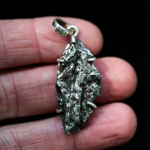 May include: A silver pendant featuring a rough, dark gray, textured meteorite. The pendant is suspended from a silver bail and secured with small silver pins. The meteorite has a unique, irregular shape, and is displayed against a dark background.