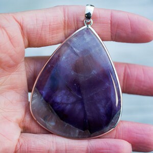 May include: A large, teardrop-shaped pendant with a polished, deep purple gemstone. The stone has lighter purple and white veining. The pendant is set in a silver-colored metal frame with a matching bail for a necklace chain. The gemstone is approximately 3 inches long.