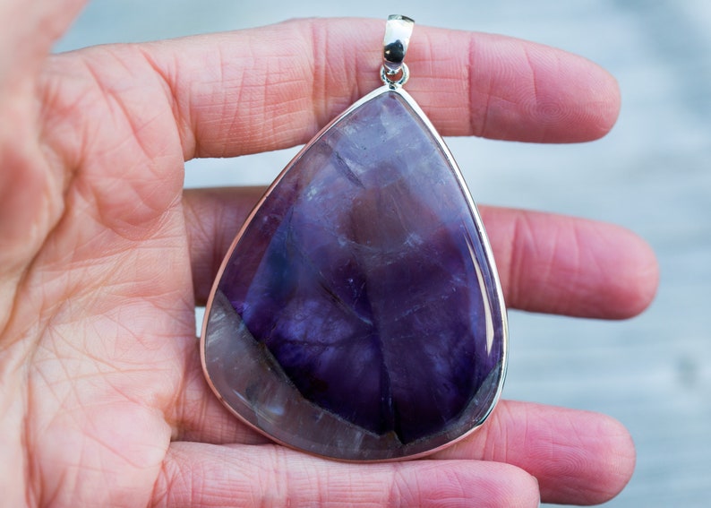May include: A large, teardrop-shaped pendant with a polished, deep purple gemstone. The stone has lighter purple and white veining. The pendant is set in a silver-colored metal frame with a matching bail for a necklace chain. The gemstone is approximately 3 inches long.