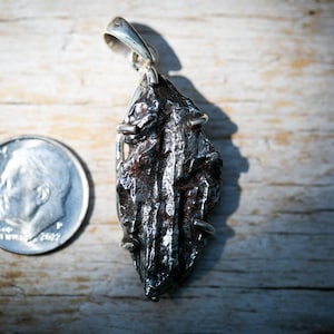 May include: A silver pendant featuring a dark, textured, irregularly shaped stone, possibly a meteorite. The pendant is suspended from a silver bail. A U.S. dime is placed next to the pendant for size comparison. The stone has a rough, metallic appearance.