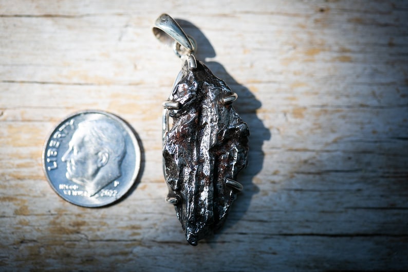 May include: A silver pendant featuring a dark, textured, irregularly shaped stone, possibly a meteorite. The pendant is suspended from a silver bail. A U.S. dime is placed next to the pendant for size comparison. The stone has a rough, metallic appearance.