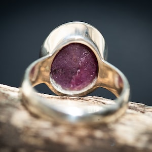May include: Close-up of a silver ring with a large, oval-shaped, deep pink gemstone. The ring's band is a polished silver, and the gemstone has a slightly textured surface. The ring is set against a dark background.