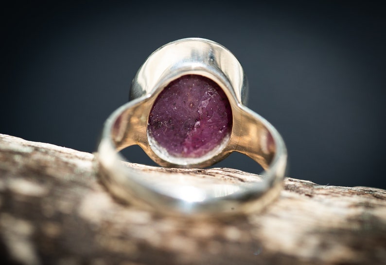 May include: Close-up of a silver ring with a large, oval-shaped, deep pink gemstone. The ring's band is a polished silver, and the gemstone has a slightly textured surface. The ring is set against a dark background.