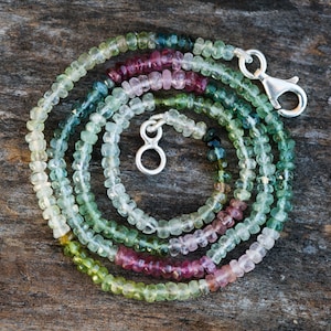 May include: A multi-coloured tourmaline gemstone necklace with a silver clasp. The necklace is made of small, faceted beads in shades of green, blue, pink, and white. The necklace is coiled on a wooden surface next to a US dime for scale.