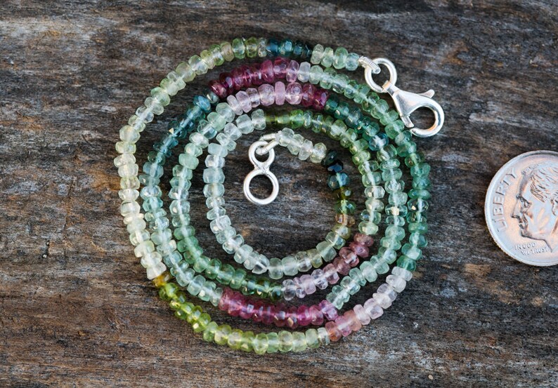 May include: A multi-coloured tourmaline gemstone necklace with a silver clasp. The necklace is made of small, faceted beads in shades of green, blue, pink, and white. The necklace is coiled on a wooden surface next to a US dime for scale.