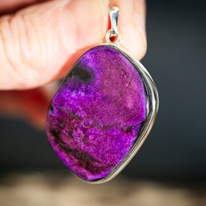 May include: A vibrant purple pendant with a silver-colored frame. The stone has a textured surface with dark purple and black veining. The pendant is held up by a silver bail.