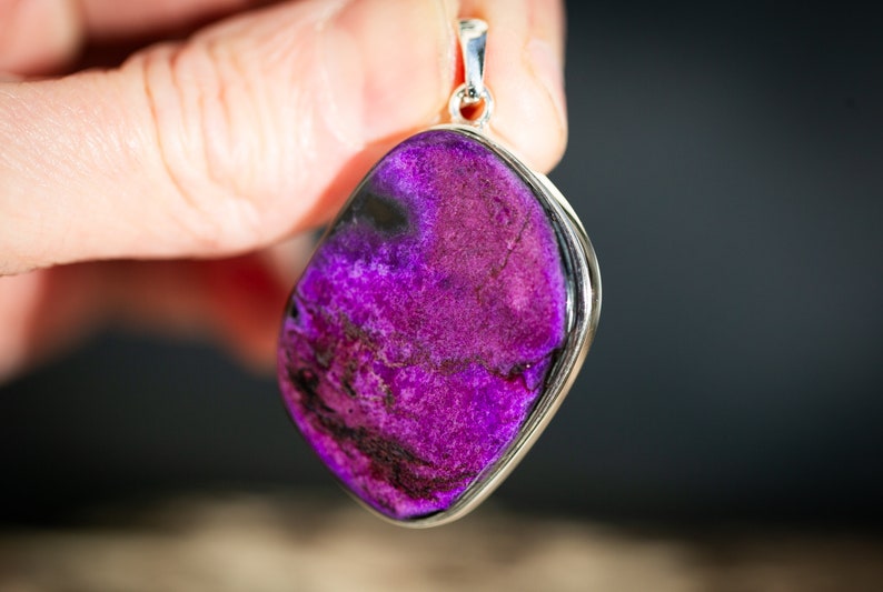 May include: A vibrant purple pendant with a silver-colored frame. The stone has a textured surface with dark purple and black veining. The pendant is held up by a silver bail.