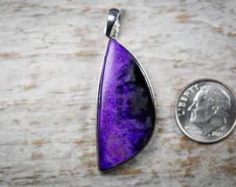 May include: A pendant with a vibrant purple and black stone, set in a silver-colored metal frame. The pendant is next to a U.S. dime coin for size comparison. The stone has a unique, marbled appearance.