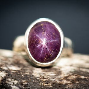 May include: A close-up of a silver ring with a large, oval-shaped, deep purple gemstone. The gemstone has a star-like pattern across its surface. The ring is set on a piece of weathered wood.