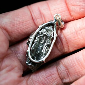 May include: A silver pendant featuring a dark, textured, irregularly shaped stone. The stone is encased in a silver setting with a loop for a chain. The pendant is held in a person's hand, with the background out of focus.